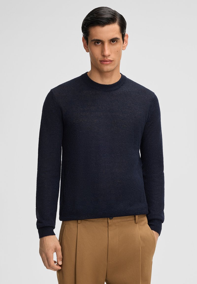 Marineblauer Rundhalsausschnitt-Pullover aus leichtem Stoff, mit langen Ärmeln und einer subtilen Textur; kombiniert mit sandfarbenen, gefalteten Hosen.