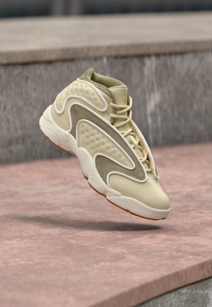 Jasnobeżowy sneaker z falowanymi białymi i szarymi nakładkami, teksturowanymi panelami oraz gumową podeszwą unoszącą się nad kamienną powierzchnią.