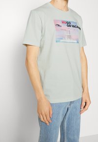 Camiseta de algodón verde claro con un diseño impreso colorido que incluye el texto "HUGO DE VACACIONES", mangas cortas y cuello redondo.