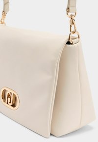 Krämfärgad handväska i fuskläder med en strukturerad form, guldfärgad metallutrustning och ett vikbart lock som stängning. Har en avtagbar axelrem.