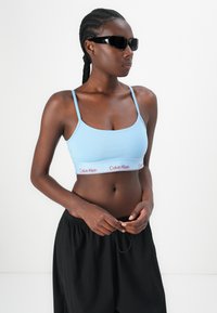 Débardeur court bleu clair avec des fines bretelles, présentant une taille blanche avec le logo rouge "Calvin Klein". Associé à un short noir ample.