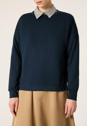Personne portant un pull bleu marine à manches longues superposé à une chemise grise à col et une jupe beige, debout devant un fond clair.