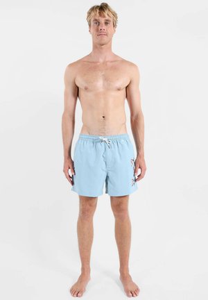Jeune homme debout pieds nus portant un short de bain bleu clair avec lien de serrage et motif floral sur la jambe droite, fond neutre.
