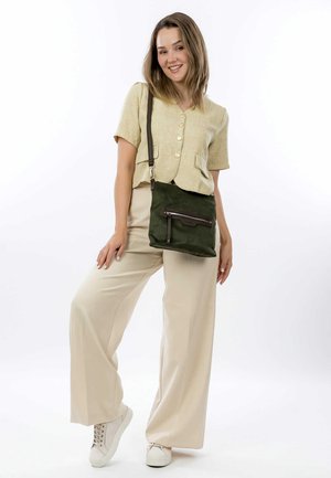 Green suede crossbody bag with a front zip pocket, adjustable strap, and textured accents. Paired with light beige wide-leg pants and a button-up top.