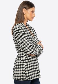 Blazer de patrón de pata de gallo en blanco y negro con un acabado texturizado, que presenta un cuello de muesca y mangas largas. Vista lateral.
