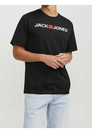 Man wearing a black Jack & Jones logo t-shirt and light blue jeans, standing with hands loosely clasped in front.