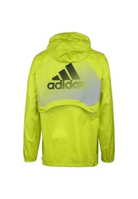 Lys gul vindjakke med hætte, der har et sort Adidas-logo og grafisk mønster på ryggen. Glat, letvægtsstof.