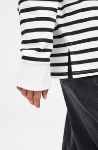 Chemise à rayures noir et blanc à manches longues avec un poignet blanc et des fentes sur les côtés, portée sur un pantalon sombre, mettant en avant un style décontracté et des couleurs contrastées.