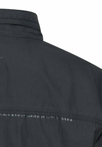 Czarna kurtka widziana od tyłu, z naciskiem na kołnierz i górną część ramienia, z widocznym przeszyciem oraz odblaskowym paskiem poniżej ramienia.