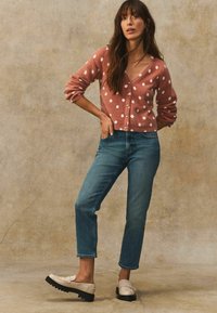 Roze polkadot cardigan, blauwe jeans en beige instappers met zwarte zolen. Textured breiwerk, V-hals en omgeslagen mouwen accentueren de outfit.