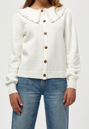 Kvinde iført hvid knappet cardigan med flæsekrave og blå denimjeans, stående med hænderne afslappet ned langs siderne.