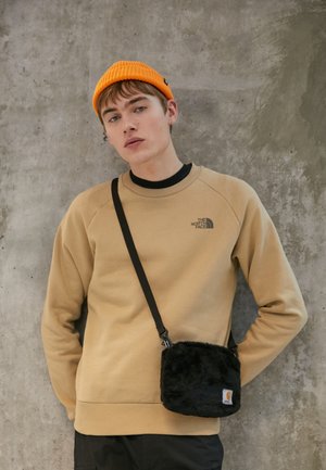 Felpa beige con logo, abbinata a una borsa in pelliccia sintetica nera con tracolla. Berretto lavorato a maglia arancione. Sfondo semplice di una parete grigia texturizzata.