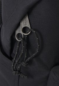 Detail černého látkového batohu se stříbrnými zipy a černými šňůrkami. Textura je hladká s jemným tkaným vzorem.