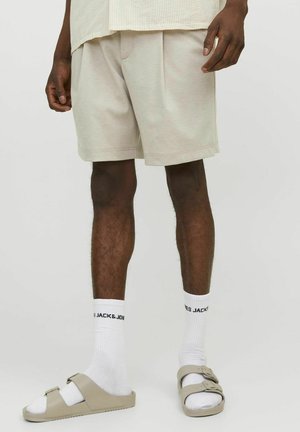 Bermudas de color beige claro con pliegues, llevadas con calcetas blancas que tienen texto negro y sandalias de color beige claro con dos tiras y una hebilla.