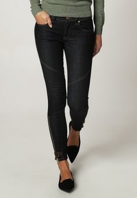 Tmavě modré džíny s úzkým střihem, které mají diagonální švy a zipy na kotnících, doplněné černými podpatky.