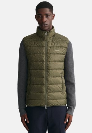 Olive green puffer vest with quilted texture, high collar, zip closure, and side pockets, worn over a gray long-sleeve top.
