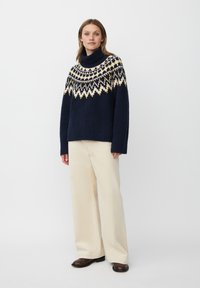 Granatowy sweter warkoczowy z wysokim kołnierzem, ozdobiony wzorami w śnieżynki w kolorze kremowym i musztardowym. Połączony z kremowymi spodniami o szerokiej nogawce i brązowymi butami.