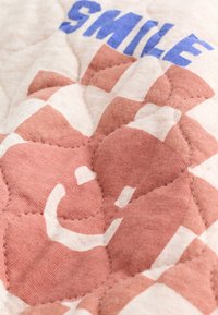 Tissu matelassé avec un visage souriant blanc imprimé et des lettres bleues en bloc formant le mot « SMILE » sur un fond rose poudré et crème.