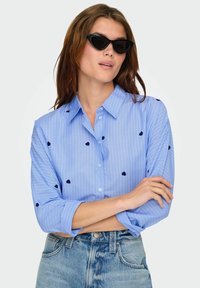 Šviesiai mėlyna marškinė su vertikaliomis baltomis juostelėmis, dekoruoti tamsiai mėlynu širdelės siuvinėjimu. Suporuoti su aukštu liemeniu džinsais.