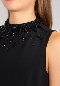 Černé bezramínkové šaty s vysokým výstřihem zdobeným černými korálky a krystaly, což vytváří texturovaný, dimenzionální efekt.