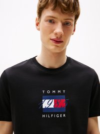 T-shirt en coton noir avec un design de logo central comportant des accents rouges, blancs et bleus, et le texte "TOMMY HILFIGER 1985" en gras.