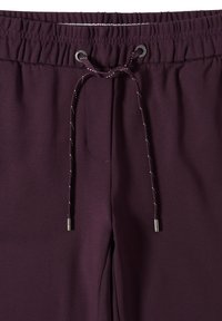 Burgundské tepláky mají pružný pás s šňůrkou, kovové očíslování a hladkou texturu se bočními kapsami.