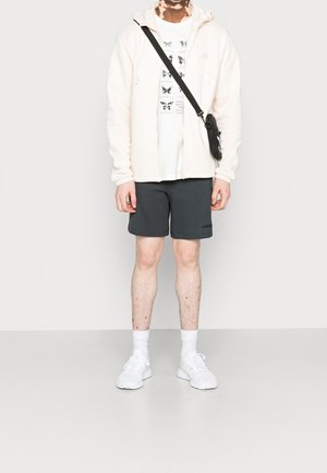Jeune portant un sweat à capuche blanc sur un t-shirt imprimé de papillon, short gris foncé, baskets blanches et sac bandoulière sur fond uni.