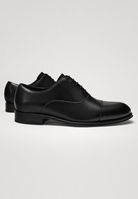 Paire de chaussures habillées Oxford en cuir noir avec laçage fermé, bout rapporté et petits talons empilés sur un fond clair.