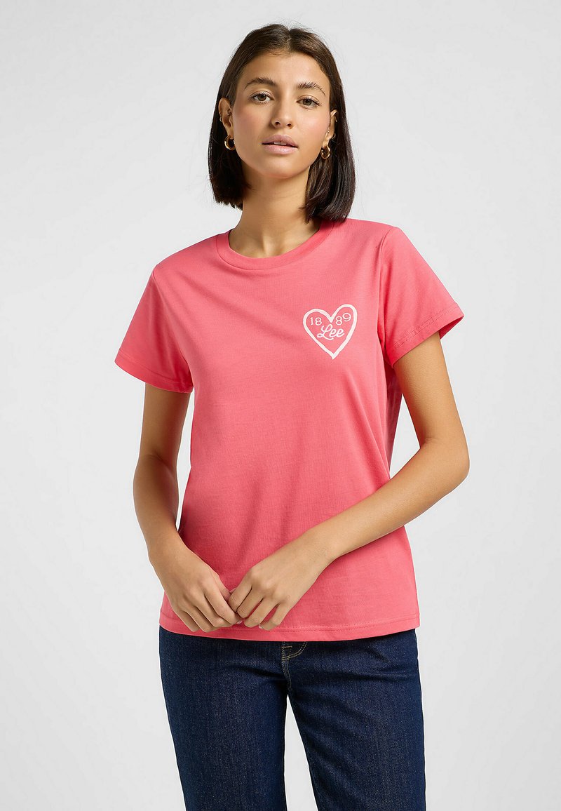 T-shirt rose à manches courtes en coton avec un col rond. Présente un graphique en forme de cœur blanc avec le texte "Lee 1889" sur la zone à gauche de la poitrine.