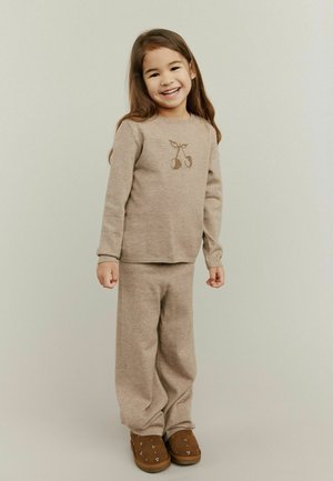 Karamelīgu adītu džemperi ar ķiršu dizainu, apvienojot to ar atbilstošām brīva piegriezuma biksēm. Brūni slip-on apavi ar dekoratīvām studām.