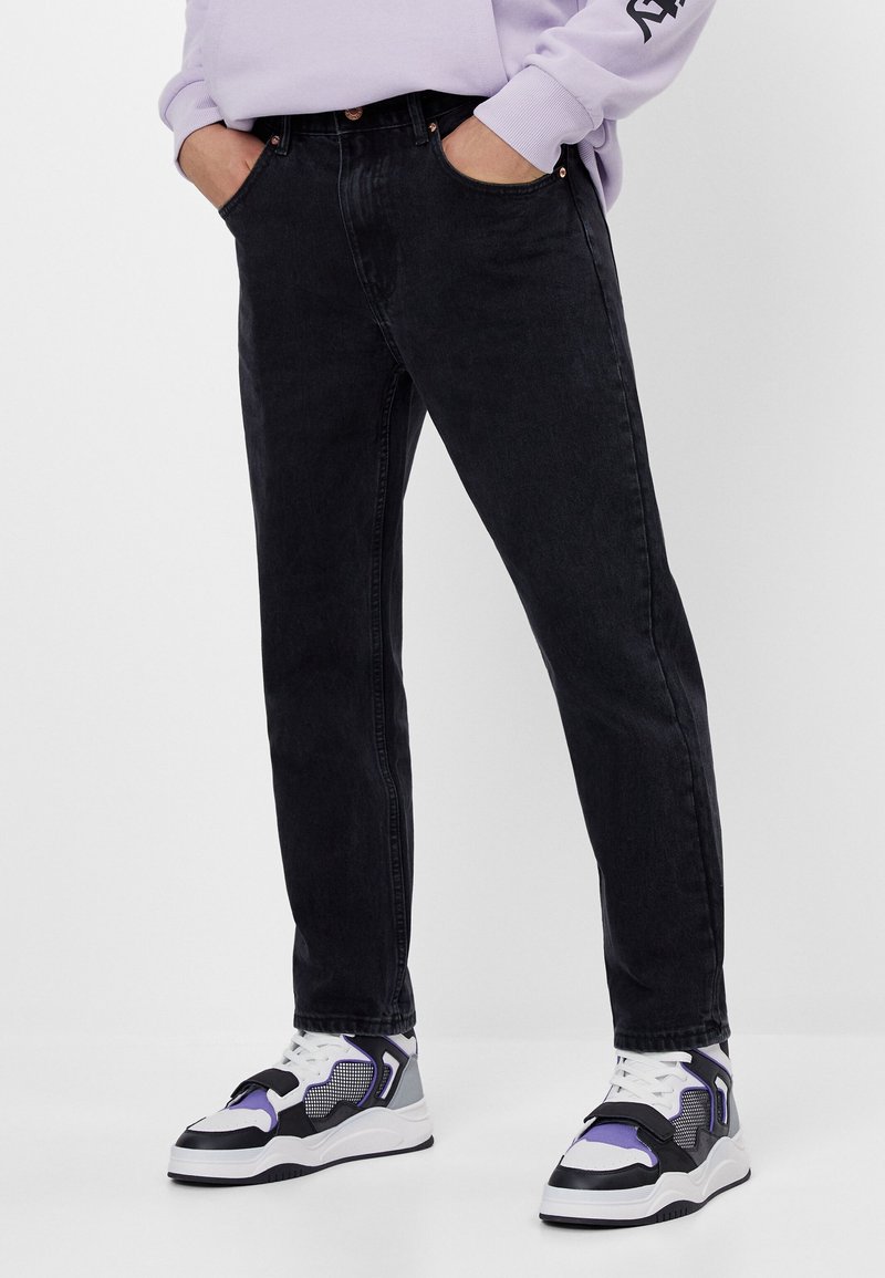 Jeans de mezclilla negra con un corte slim, con cinco bolsillos y costuras visibles. Combinados con zapatillas deportivas blancas y moradas.