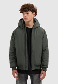 Jongere met krullend haar, gekleed in een donkergroene hoodie over een zwart shirt, staand tegen een effen lichte achtergrond.