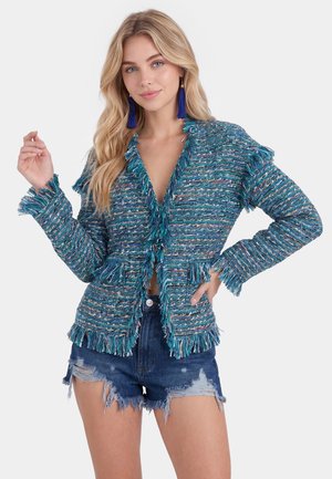 Blonde Frau trägt eine blau-grüne, strukturierte Fransenjacke mit blauen Quastenohrringen und ausgewaschenen Jeansshorts, posiert mit der Hand in der Hüfte.