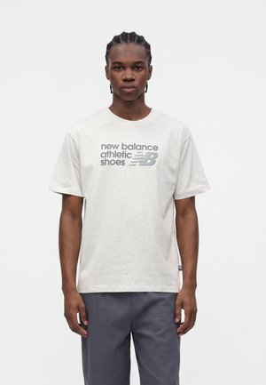 Młody mężczyzna z warkoczykami, ubrany w jasnoszary t-shirt z logo New Balance i ciemnoszare spodnie, stojący na białym tle.