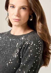Grauer Strickpullover mit kurzen Ärmeln, verziert mit silbernen Pailletten auf der Vorderseite. Glatte Textur, runder Ausschnitt und figurbetontes Design.