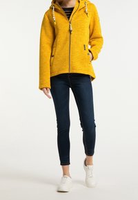 Osoba v žluté zipové mikině, tmavých skinny džínách a bílých teniskách, stojící před čistým bílým pozadím.