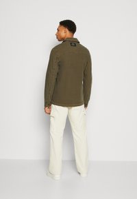 Chaqueta de camisa de color verde olivo con corte recto, puños abotonados y etiqueta en la parte posterior, combinada con pantalones cargo blancos. Textura suave.
