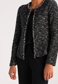 Veste texturée noir et blanc avec un design court, un col rond et des bords effilochés. Présente un devant sans boutons et des manches longues.