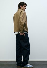 Mladý muž v béžové bundě, pruhovaném tričku, volných tmavých džínách a modrých teniskách stojí s rukou v zadní kapse opřený o jednobarevnou zeď.