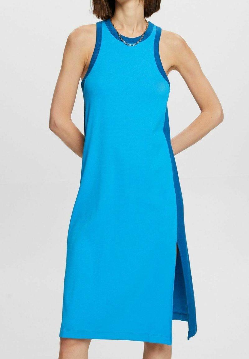 Femme portant une robe bleue sans manches jusqu’aux genoux avec un bord bleu foncé et une fente latérale, debout, les mains derrière le dos.