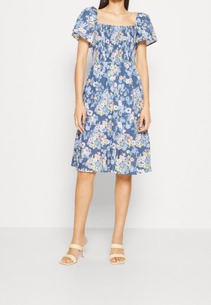 Žena v modrých květovaných šatech po kolena s nafouklými rukávy a hranatým výstřihem, doplněná béžovými sandály na podpatku.