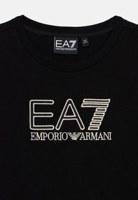 Melna kokvilnas T-kreklu ar apaļu kakla izgriezumu. Izteiksmīgs balts izšūts logos: "EA7 EMPORIO ARMANI" ar stilizētiem elementiem un akcentiem.