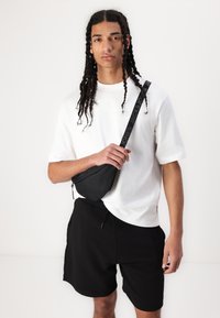 Čierna crossbody taška s textúrovaným povrchom, popruhom s logom a zipsom, nosená cez biely tričko a čierne šortky.