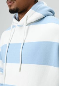 Sweat à capuche présentant des rayures alternées bleu clair et blanc. Confectionné en tissu doux avec des cordons de serrage et un col décontracté.