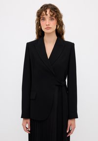 Černý blazer s překládaným designem, dlouhými rukávy a dvěma předními kapsami. Vyroben z hladké látky, s uvázaným detailem v pase.