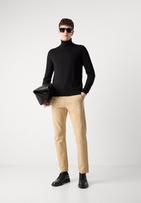 Cămașă neagră tricotată cu guler înalt, pantaloni bej deschis și pantofi din piele negri. Ținând o geantă structurat neagră cu un finisaj mat.