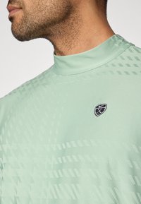 Πράσινο πουκάμισο κοντομάνικο μέντας με ψηλό γιακά. Διαθέτει γεωμετρικό υφασμάτινο σχέδιο και μικρό μαύρο λογότυπο στο αριστερό στήθος.