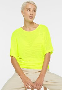 Neon-gelbes Strickoberteil mit kurzen Ärmeln, lässiger Passform und strukturiertem Muster. Kombiniert mit hellbeigen Hosen, während im Sitzen getragen.