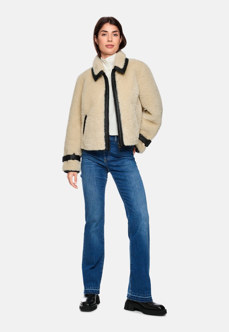 Beige shearling jacket with black trim, zip front, and side pockets, paired with blue flared jeans and black ankle boots.