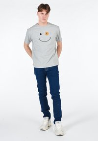 T-shirt in cotone grigia con un design di faccina sorridente con un accento giallo, abbinata a jeans blu scuro e sneakers bianche.